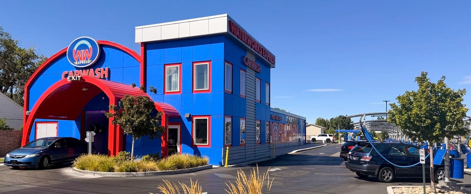 The WaterWorks Express car wash on 7800 South in West Jordan on a clear day: a car leaving the red arched tunnel exit, with the row of free self-serve vacuum stalls in use on the right.
